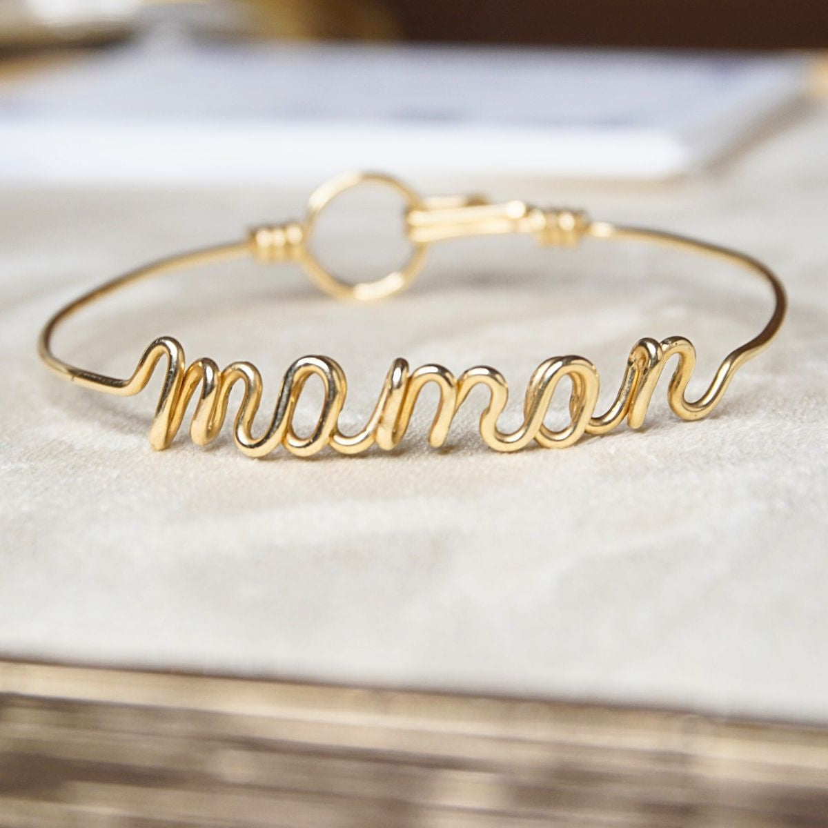 Bracelet prénom personnalisé en fil doré, formé à la main, sur fond beige doux avec mise au point sur le bijou.